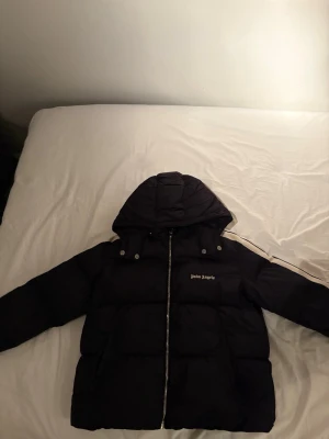 Svart pufferjacka från Palm Angels - Svart pufferjacka från Palm Angels med huva och silverfärgad dragkedja framtill. Jackan har en quiltad design och logotyp på bröstet. Perfekt för kalla dagar med sin tjocka och vadderade look. Klassisk passform och långärmad modell.