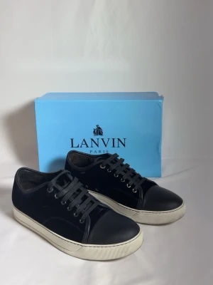 Svarta sneakers från Lanvin - Stilrena svarta sneakers från Lanvin med rund tå, vita platta sulor och svarta snören. Ovandelen är i en mix av skinn och syntetmaterial med diskreta sömdetaljer. Perfekta för dig som gillar en clean och klassisk look.