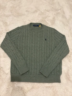 Grå kabelstickad tröja från Polo Ralph Lauren - Kabelstickad tröja i ljusgrått från Polo Ralph Lauren med rund halsringning och klassisk marinblå logga broderad på bröstet. Tröjan har långa ärmar och ribbade muddar vid ärmslut och nederkant. Perfekt för lager på lager och stilren vardagslook.