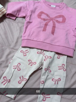 Rosa set med rosetttryck - Sött tvådelat set med rosa sweatshirt och matchande byxor. Tröjan är ljusrosa med stor rosett framtill, byxorna är vita med rosa rosetter över hela. Perfekt för dig som gillar gulliga detaljer och mjuka material.