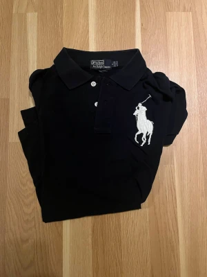 Svart pikétröja från Ralph Lauren - Klassisk mörkblå pikétröja från Ralph Lauren med vit broderad logga på bröstet. Tröjan har krage, knappar framtill och korta ärmar. Perfekt för en stilren och sportig look. Är Small men passar om man har M