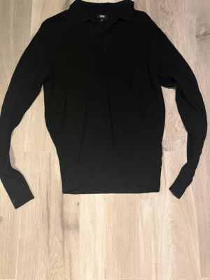 Svart pikétröja från Uniqlo - Klassisk svart långärmad pikétröja från Uniqlo i mjukt stickat material. Tröjan har krage, knäppning framtill och ribbade muddar vid ärmslut och nederkant. Perfekt för en clean och stilren look.