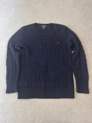Mörkblå kabelstickad tröja Polo Ralph Lauren - Kabelstickad mörkblå tröja från Polo Ralph Lauren med klassisk röd logga broderad på bröstet. Rund halsringning och ribbade muddar vid ärmslut och nederkant. Perfekt till lager på lager under höst och vinter.