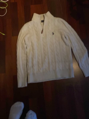 Vit kabelstickad tröja Polo Ralph Lauren - Kabelstickad vit tröja från Polo Ralph Lauren med halv dragkedja och klassisk marinblå logga på bröstet. Tröjan har ribbade muddar och hög krage, perfekt för lager på lager. Snygg och tidlös design som passar till det mesta.