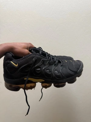 Nike Air Vapormax Plus svart/guld - Nike Air Vapormax Plus sneakers i svart med snygga gulddetaljer och ikonisk synlig sula. Ovandelen är i syntetmaterial med vågformade detaljer och snörning. Perfekta för dig som vill ha en sportig och modern look.