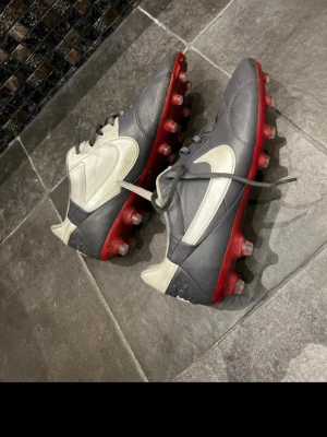 Nike grå-vita fotbollsskor med röd sula - Snygga fotbollsskor från Nike i grått och vitt med klassisk swoosh på sidan. Skorna har snörning och en röd sula med dobbar för bra grepp på planen. Tillverkade i syntetmaterial och designade för att ge stabilitet och komfort under matchen. Lite smutsiga på bilden men knappt använda så det borde gå att tvätta, min brorsas gamla som han knappt använt 