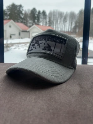 Grå keps med hockeymotiv - Säljer en grå keps med böjd skärm och mesh på sidorna. Framtill finns ett svartvitt patch med hockeymotiv och texten 'Tiaris'. Kepsen har klassisk trucker-modell och justerbar baksida för skön passform. Perfekt för dig som gillar sportig stil.