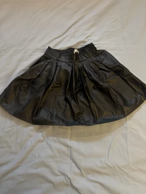 Svart kortkjol i skinnimitation - Säljer en svart kortkjol i skinnimitation med resår i midjan. Kjolen har en lätt utställd form och ger en cool vibe till din outfit. Perfekt att matcha med en oversized tröja eller crop top.