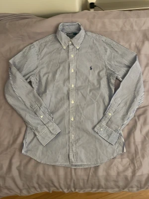 Randig skjorta från Polo Ralph Lauren - Klassisk skjorta från Polo Ralph Lauren med smala blå och vita ränder. Skjortan har button-down krage, långa ärmar och broderad logga på bröstet. Tillverkad i bomull och stängs med vita knappar framtill. Kan gå ner i pris vid snabb försäljning!