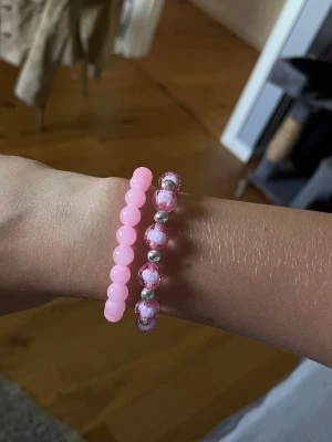 Två rosa pärlarmband - Säljer två söta armband med pärlor i olika nyanser av rosa. Det ena armbandet har enfärgade ljusrosa pärlor medan det andra har rosa och silverfärgade pärlor i ett mönster. Båda är elastiska och passar de flesta handleder. Perfekt för att addera lite färg till din stil!