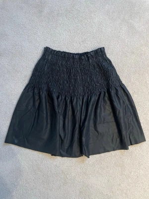 Svart ”läder” kortkjol från Zara - Säljer en svart ”läder” kortkjol från Zara med smockad midja och lätt utställd passform. Kjolen är enkel och stilren, perfekt att matcha med olika toppar. Tillverkad i mjukt material som känns bekvämt hela dagen.