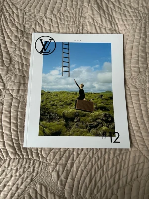 Louis Vuitton The Book #12 - Snygg och modern bok från Louis Vuitton, nummer 12 i serien 'The Book'. Omslaget har ett konstnärligt foto med en person, resväska och stege mot blå himmel och grön natur. Boken har vit ram och tryckt LV-logga. Perfekt för dig som gillar mode och inspirerande bilder.