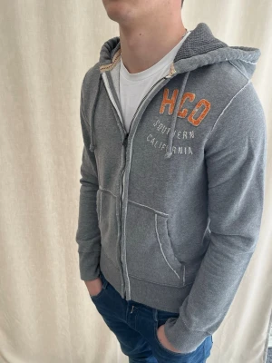 Grå zip hoodie från Hollister - Vintage design hoodie från hollister. Helt ny aldrig använd. Broderad HCO-logga. Strl S. Personen på bilden är 184 cm. Skicka prisförslag. Kolla in min profil för liknande plagg