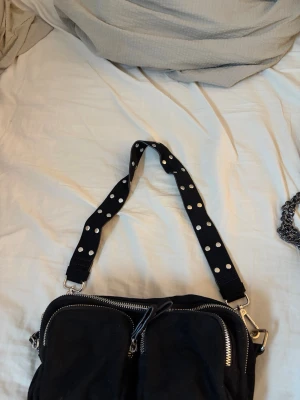 Svart axelväska med prickigt band - Snygg svart axelväska med bred axelrem i svart med vita prickar. Väskan har två stora fack framtill med silverfärgade dragkedjor och detaljer i metall. Perfekt för dig som vill ha en cool och praktisk väska till vardags.