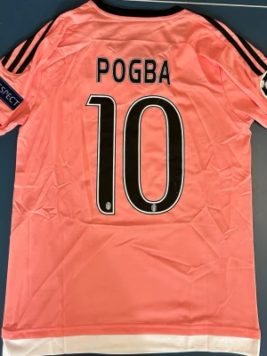 Juventus Fotbollströja Pogba #10 - Juventus matchtröja i rosa med Pogba och nummer 10 på ryggen. Jeep som sponsor och flera klubbmärken på bröstet. Svarta detaljer och ränder på axlarna, samt Champions League-märke på ärmen. Storlek M. Helt ny.
