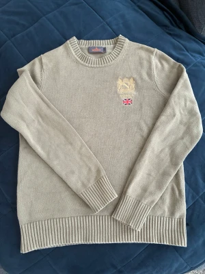 Olivgrön stickad tröja från Morris - Snygg olivgrön (färg olive) stickad tröja från Morris (Trenton oneck) med broderad krona och brittisk flagga på bröstet. Tröjan har rund halsringning, ribbade muddar och nederkant.
