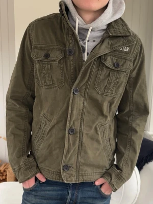 Olivgrön fieldjacket från Hollister - Snygg olivgrön fieldjacket från Hollister med fyra fickor framtill och tydliga sömmar. Jackan har knäppning med stora knappar och en klassisk krage. Perfekt lager-på-lager och passar till jeans för en avslappnad look.