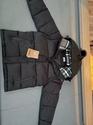 Svart dunjacka med huva från Burberry - Svart dunjacka från Burberry med huva och rutigt foder. Jackan har dragkedja framtill, tryckknappar och två sidofickor med dragkedja. Klassisk pufferstil som håller dig varm under kalla dagar. Kan även transformeras till en väst.