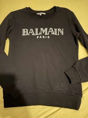 Svart sweatshirt från Balmain Paris - Svart sweatshirt från Balmain Paris med vit logotyptryck på bröstet. Tröjan har rund halsringning, ribbade muddar vid ärmslut och nederkant. Perfekt för en trendig och avslappnad stil. STORLEK M MEN SITTER SOM S!!!!!!