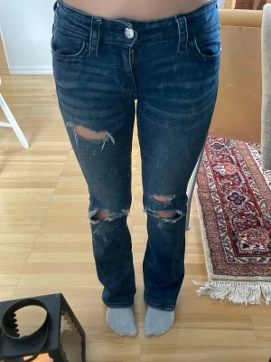 Mörkblå slitna bootcut jeans - Säljer ett par mörkblå jeans med slitningar och hål på knäna. Modellen har bootcut och låg midja, med klassiska fickor och knappdetaljer bak. Perfekta för en avslappnad och trendig look.