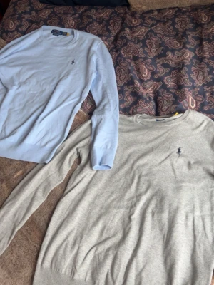 Två stickade tröjor från Polo Ralph Lauren - Säljer två stilrena stickade tröjor från Polo Ralph Lauren. En i ljusblått och en i ljusgrått, båda med den klassiska broderade loggan på bröstet. Tröjorna har rund halsringning och ribbade muddar vid ärmslut och nederkant. Perfekta för lager på lager.