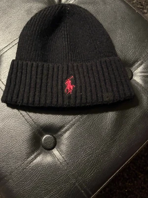 Svart ribbstickad mössa Polo Ralph Lauren - Svart ribbstickad mössa från Polo Ralph Lauren med klassisk uppvikt kant och röd broderad logga framtill. Perfekt accessoar för kyliga dagar och enkel att matcha med olika outfits. Tidlös design som funkar till det mesta.