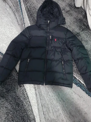 Svart dunjacka från Polo Ralph Lauren - Snygg svart dunjacka från Polo Ralph Lauren med huva och dragkedja framtill. Jackan har två snedställda fickor med dragkedja och den klassiska röda loggan på bröstet. Använt den bara 2 gånger så helt ny.Perfekt för kalla dagar och enkel att matcha med allt.