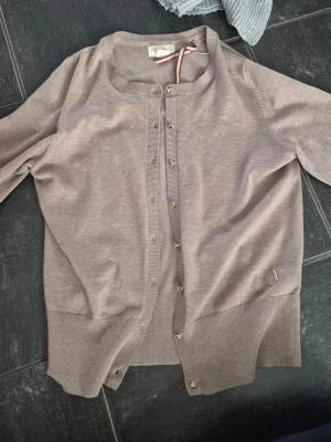 Beige kofta - Klassisk beige kofta från med rund halsringning och ribbade muddar. Koftan har knappar framtill och en liten metallbricka vid nederkanten. Perfekt att slänga över en t-shirt eller linne för en enkel och stilren look.