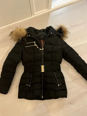 Hollies kappa med äkta päls - Snygg svart dunjacka från Hollies med guldfärgade dragkedjor, avtagbar huva med fpäls och bälte i midjan. Pyttelitet hål framtill vid dragkedjan (bild 4). Passar även 36,38. Väldigt varm under vintern och är figurnära. 
