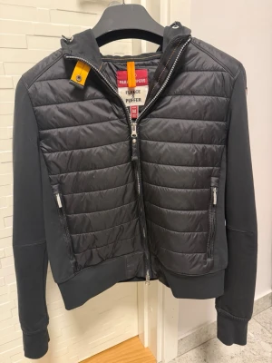 Svart pufferjacka från Parajumpers - Svart pufferjacka från Parajumpers med quiltad front och rygg, huva och dragkedja. Jackan har ribbade muddar och två fickor med dragkedja. Materialmix med polyester och bomull ger en sportig och modern look.
