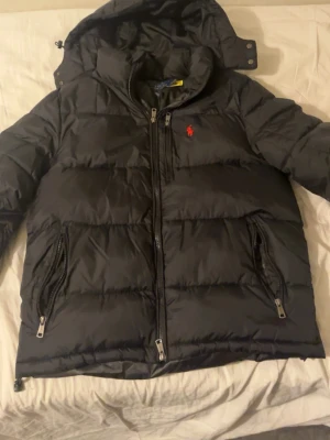 Svart dunjacka från Polo Ralph Lauren - Svart puffer dunjacka från Polo Ralph Lauren med röd broderad logga på bröstet. Jackan har huva, dragkedja framtill och två sidofickor med dragkedja. Klassisk quiltad design och varm vaddering, perfekt för kalla dagar.