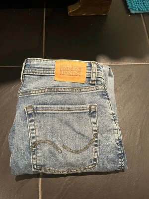 Blå jeans från Jack & Jones, stl 158 - Snygga blå jeans från Jack & Jones i klassisk modell. Jeansen har en ljus tvätt, raka ben och detaljerade sömmar på bakfickan. Perfekta för dig som gillar en enkel och tidlös stil. Märkespatch i läder baktill i midjan.