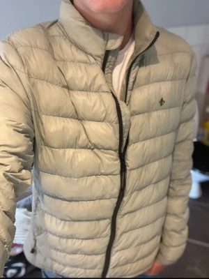 Ljusgrön dunjacka från Morris - Snygg ljusbeige dunjacka från Morris med quiltad design och hög krage. Jackan har dragkedja framtill och diskret logga på bröstet. Perfekt för kyliga dagar och enkel att matcha med olika outfits.