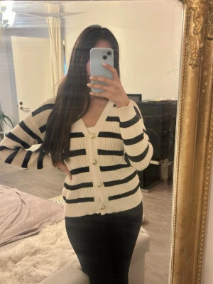 Randig kofta med pärlknappar - Snygg ribbstickad kofta från krämvit med svarta horisontella ränder. Koftan har v-ringning och pärlknappar framtill. Perfekt att slänga över en t-shirt eller linne för en trendig look.