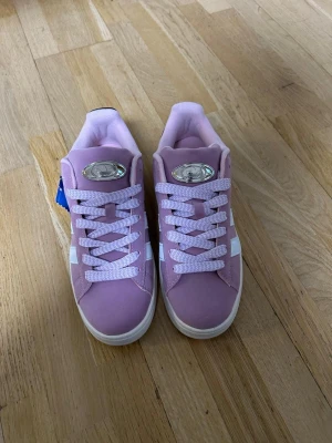 Ljuslila Adidas Campus sneakers - Säljer ett par ljuslila Adidas Campus sneakers med klassiska vita ränder på sidan och vit sula. Skorna har snörning, rund tå och är tillverkade i mocka med detaljer i syntet. Perfekta för dig som vill sticka ut med färg på fötterna.