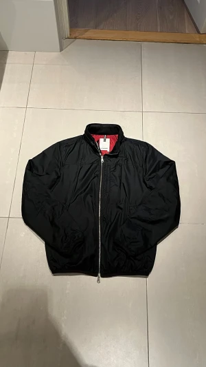 Svart bomberjacka J.Lindeberg - Snygg svart bomberjacka från J.Lindeberg med klassisk passform och silvrig dragkedja framtill. Jackan har en röd insida som ger en cool kontrast och står ut. Perfekt för dig som vill ha en stilren och trendig look.