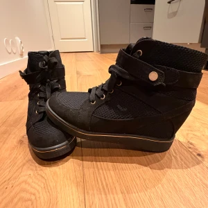 Svarta höga sneakers med snörning - Svarta sneakers med högt skaft, snörning och dekorativ rem med knappdetalj. Ovandel i textil och syntet, platt sula och rund tå. Snygga detaljer i guld vid snörningen och en diskret rand längs sulan.