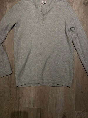 Ljusgrå henleytröja från Celio - Säljer en ljusgrå långärmad henleytröja från Celio med tre knappar vid halsen. Tröjan har en enkel och stilren design, perfekt för lager på lager och chill vardagsstil. Tillverkad i mjukt material för extra komfort.