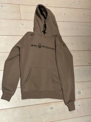 Beige hoodie från Sail Racing - Snygg beige hoodie från Sail Racing med svart logga på bröstet. Klassisk känguruficka framtill och huva med snörning. Tillverkad i mjuk bomullsmix som känns skön mot huden. Perfekt för en chill och avslappnad stil.