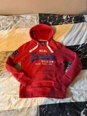 Röd Superdry hoodie - Snygg röd hoodie från Superdry med stor logga och japansk text på bröstet. Hoodien har vita dragsnören, känguruficka och ribbade muddar. Detaljerad brodyr på ena ärmen och klassisk huva. Perfekt för en avslappnad streetwear-look. Priset är diskuterbart 