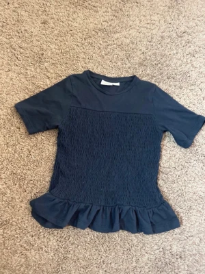 Mörkblå topp med volangkant - Snygg mörkblå topp med korta ärmar och smockad mittdel. Toppen har en volangkant nedtill som ger en extra touch. Perfekt att styla med jeans eller kjol för en trendig look.