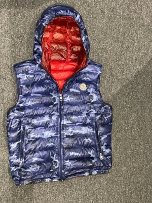 Blå camo dunväst från Moncler - Snygg blå camo dunväst från Moncler med röd insida och huva. Västen har två dragkedjefickor framtill och en Moncler-logga på bröstet. Perfekt för lager-på-lager och ger en cool streetwear-look. Tillverkad i äkta dun för extra värme.