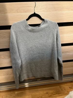 Grå stickad tröja med rund hals - En enkel och stilren grå stickad tröja med rund halsringning och långa ärmar. Tröjan har ribbade muddar vid ärmslut och nederkant. Perfekt för lager på lager och passar till det mesta i garderoben.