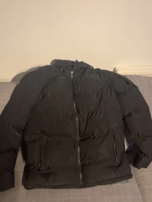 Svart pufferjacka med huva - Säljer en svart pufferjacka med huva och dragkedja framtill. Jackan har två stora fickor med dragkedja och en klassisk quiltad look. Perfekt för kalla dagar och riktigt skön passform. Passar dig som gillar en enkel och stilren vinterjacka.