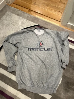 Grå sweatshirt från Moncler - Snygg grå sweatshirt från Moncler med broderad logga och text på bröstet i blått och rött. Tröjan har rund halsringning, ribbade muddar vid ärmslut och nederkant samt en avslappnad passform. Perfekt för en casual look.