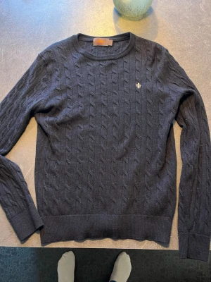 Mörkblå kabelstickad tröja Morris - Snygg mörkblå kabelstickad tröja från Morris med rund halsringning och diskret broderad logga på bröstet. Tröjan har långa ärmar och ribbade muddar vid ärmslut och nederkant. Perfekt för en stilren och klassisk look. Använd ett fåtal gånger .