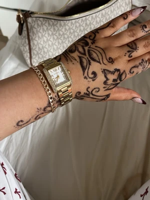 Guldfärgad klocka Michael Kors - Snygg guldfärgad klocka från Michael Kors med rektangulär urtavla, romerska siffror och gnistrande stenar runt boetten. Länkar i metall ger en lyxig känsla och klockan har en klassisk, elegant design som passar till många stilar. Original pris - 2200kr