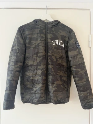 Grön camouflagejacka från SVEA - Snygg camouflagefärgad dunjacka från SVEA med huva och dragkedja framtill. Jackan har ett broderat SVEA-tryck på bröstet och en patch på ärmen. Perfekt för kalla dagar och ger en cool streetwear-look.