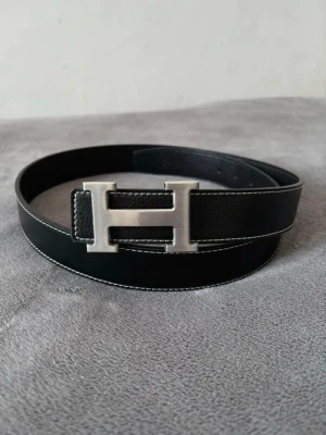 Svart läderbälte med H-spänne - Snyggt svart bälte i läder med ett stort, silverfärgat H-format spänne. Bältet har vit kontrastsöm längs kanterna och en stilren, klassisk design som passar till jeans eller kostymbyxor. Perfekt accessoar för dig som gillar exklusiv och tidlös stil.