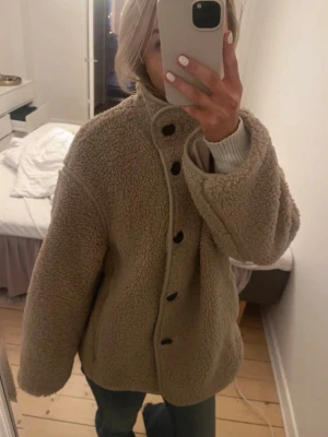Beige teddyjacka med stora knappar - Mysig beige teddyjacka med oversized passform och stora svarta knappar framtill. Jackan har hög krage och långa ärmar, perfekt för kyliga dagar. Tillverkad i mjukt teddy-material som ger en trendig och avslappnad look.